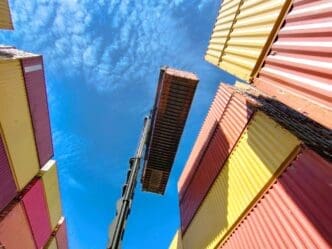 A dramatic low-angle shot from a shipping port, looking up at a crane lifting a single red container against a bright blue sky with white clouds. Stacks of yellow, red, and beige containers surround the central action.