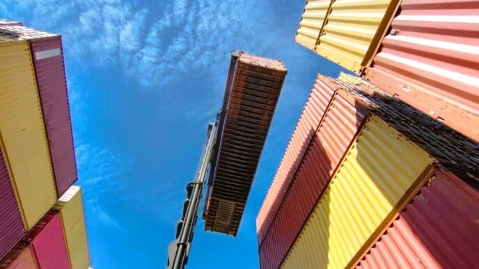 A dramatic low-angle shot from a shipping port, looking up at a crane lifting a single red container against a bright blue sky with white clouds. Stacks of yellow, red, and beige containers surround the central action.