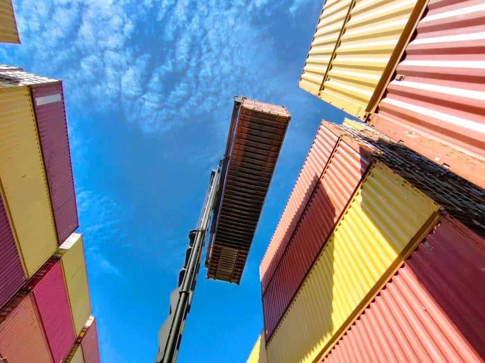 A dramatic low-angle shot from a shipping port, looking up at a crane lifting a single red container against a bright blue sky with white clouds. Stacks of yellow, red, and beige containers surround the central action.