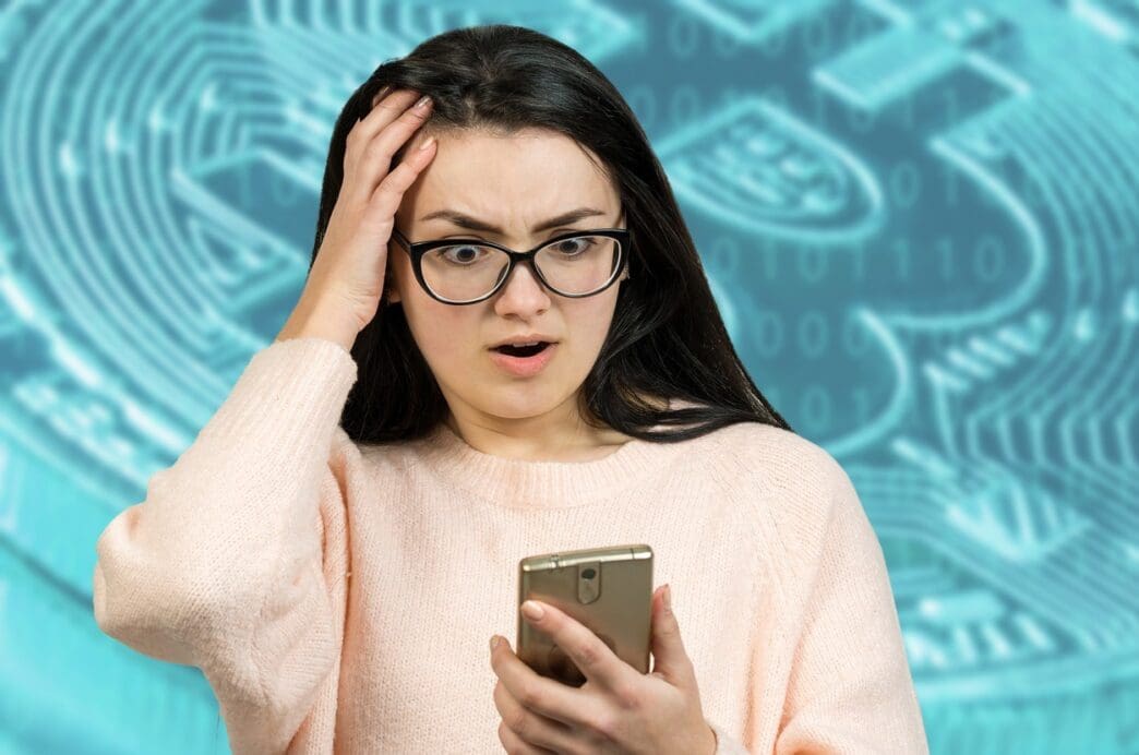 A young woman in glasses looks shocked and holds her head while looking at her phone