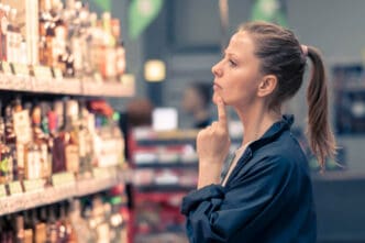 A woman with a thoughtful expression stands in a store aisle, looking at a shelf of bottles and holding her finger to her chin.