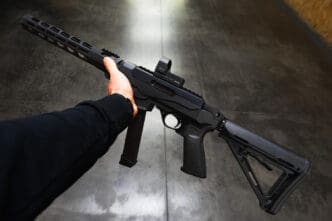 A first-person perspective of a person's hand holding a short-barreled black rifle with a red dot sight on top.
