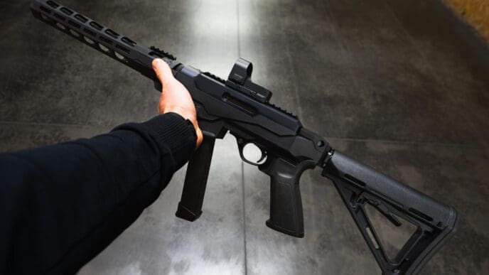 A first-person perspective of a person's hand holding a short-barreled black rifle with a red dot sight on top.