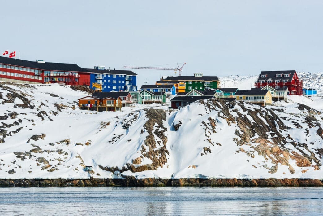 Colorful houses stand on a snow-covered rocky hillside next to a body of water in Sisimiut, Greenland