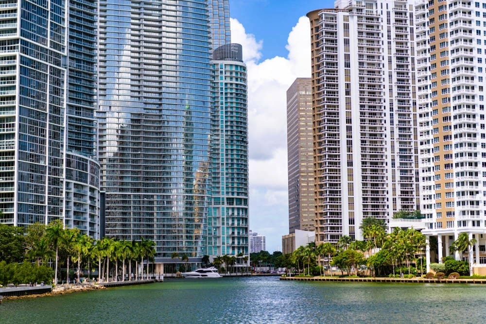 The dense and modern skyline of Brickell Key, Miami, with tall glass and white skyscrapers rising above a waterway where a luxury yacht is docked, and a line of palm trees below, under a blue sky with white clouds.