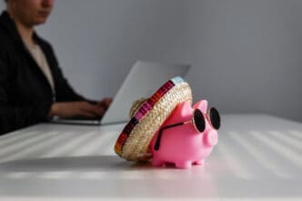 A pink piggy bank wearing a sombrero and sunglasses on a desk, with a person working on a laptop in the blurred background.