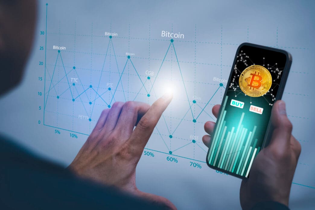 A person's hands are interacting with a smartphone displaying a Bitcoin trading app and a larger holographic-style screen with financial charts.