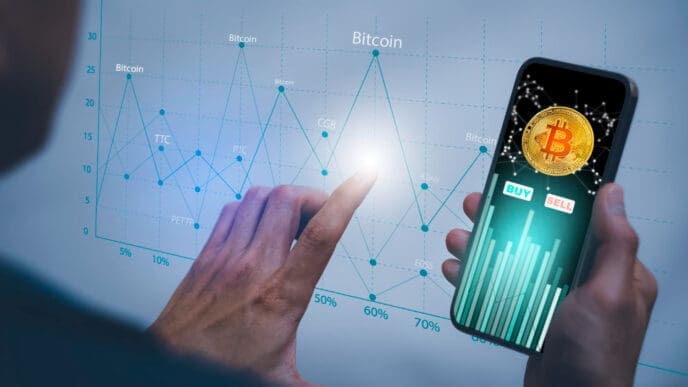 A person's hands are interacting with a smartphone displaying a Bitcoin trading app and a larger holographic-style screen with financial charts.