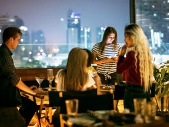 A group of young friends socializes on a rooftop balcony at night, with a city skyline in the background.