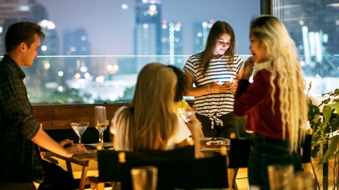 A group of young friends socializes on a rooftop balcony at night, with a city skyline in the background.