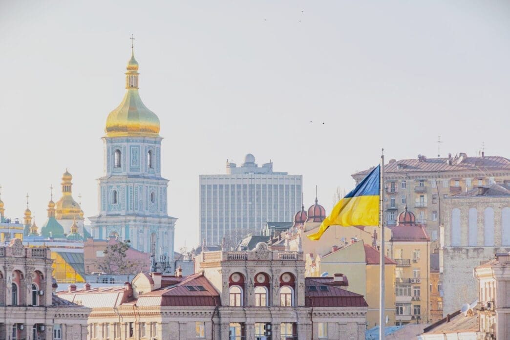 The Ukrainian flag flies in front of the skyline of Kyiv, with the golden domes and tower of St. Sophia's Cathedral in the background.