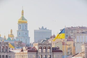 The Ukrainian flag flies in front of the skyline of Kyiv, with the golden domes and tower of St. Sophia's Cathedral in the background.