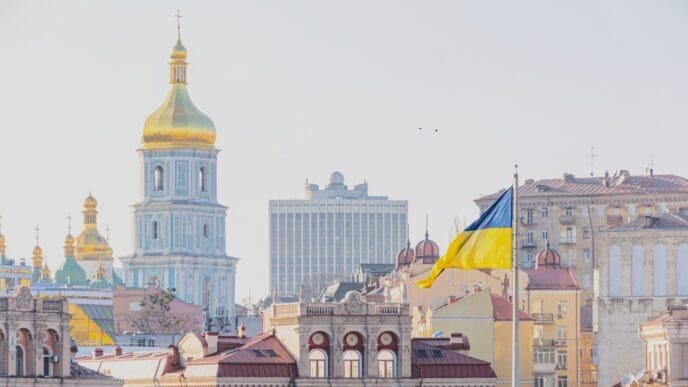 The Ukrainian flag flies in front of the skyline of Kyiv, with the golden domes and tower of St. Sophia's Cathedral in the background.