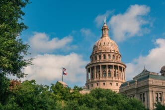 The distinctive dome and facade of the Texas State Capitol building are partially visible above a canopy of green trees.