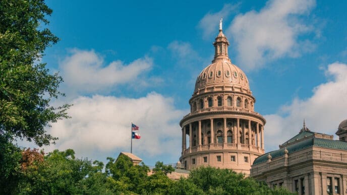 The distinctive dome and facade of the Texas State Capitol building are partially visible above a canopy of green trees.