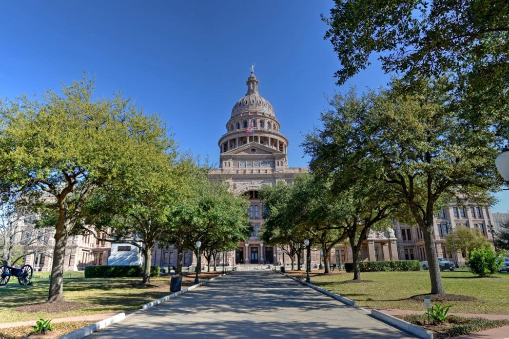 The Texas State Capitol building with its dome and a statue on top is visible from the end of a long walkway lined with trees.