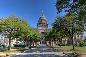The Texas State Capitol building with its dome and a statue on top is visible from the end of a long walkway lined with trees.