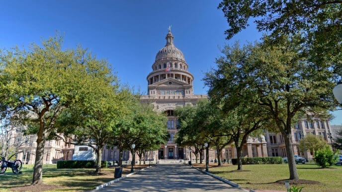 The Texas State Capitol building with its dome and a statue on top is visible from the end of a long walkway lined with trees.