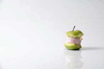 An eaten green apple core stands on a white, reflective surface, with a pink measuring tape wrapped around it, symbolizing the concept of dieting and a "detox myth."