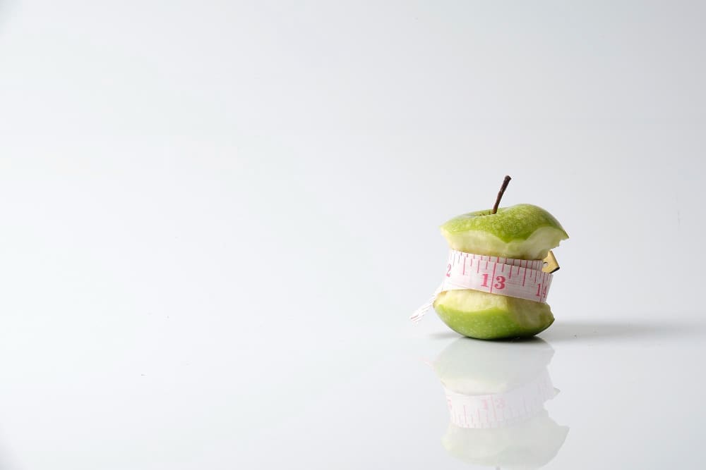 An eaten green apple core stands on a white, reflective surface, with a pink measuring tape wrapped around it, symbolizing the concept of dieting and a "detox myth."