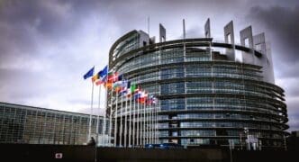 The modern, circular building of the European Parliament in Strasbourg, with a row of European flags flying in front of it under a cloudy sky.