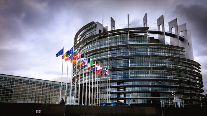 The modern, circular building of the European Parliament in Strasbourg, with a row of European flags flying in front of it under a cloudy sky.