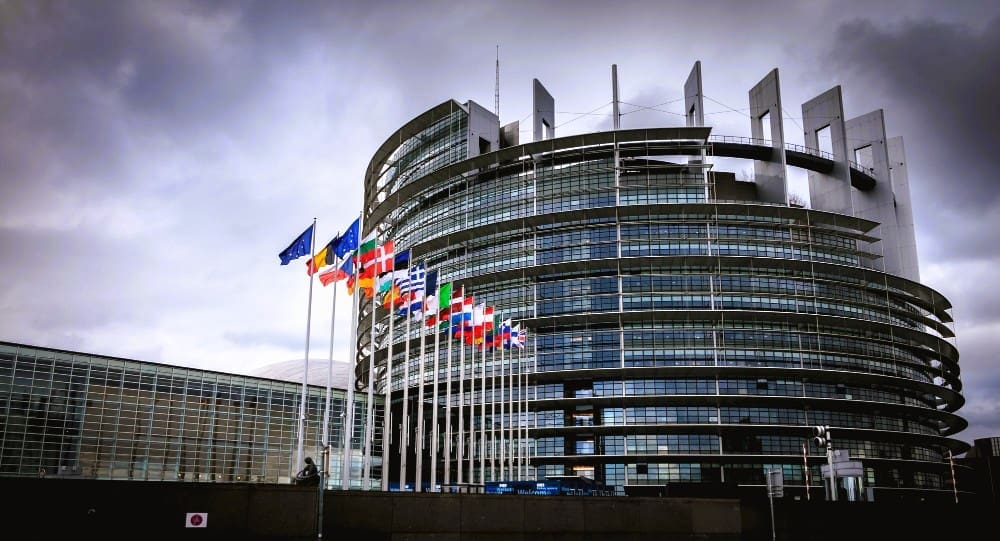 The modern, circular building of the European Parliament in Strasbourg, with a row of European flags flying in front of it under a cloudy sky.