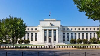 The white, neoclassical facade of the Federal Reserve Building is visible from across a street on a sunny day with a clear blue sky.