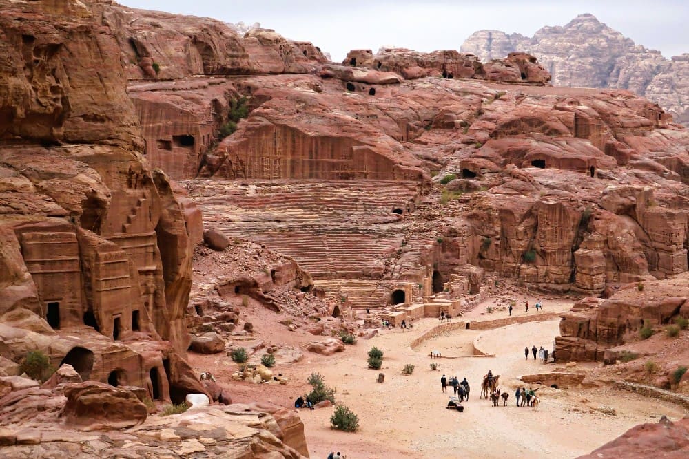 A wide view of the ancient rock-carved theater and tombs in Petra, Jordan, with tourists and camels on the desert floor.