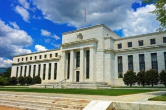 A wide view of the Marriner S. Eccles Federal Reserve Board Building, a large white neoclassical structure with columns and a grand staircase, under a bright blue sky with clouds.