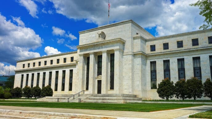 A wide view of the Marriner S. Eccles Federal Reserve Board Building, a large white neoclassical structure with columns and a grand staircase, under a bright blue sky with clouds.