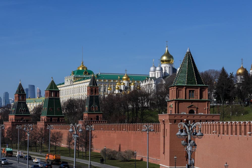 The Moscow Kremlin and its red walls, green-roofed towers, and golden domes are seen from across a busy road on a clear day.