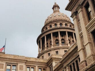 An architectural shot of the Texas State Capitol building, featuring its grand dome and columns, with a US flag at half-staff in the foreground, under an overcast sky.