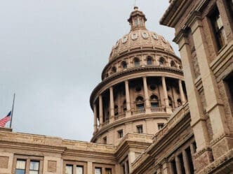 An architectural shot of the Texas State Capitol building, featuring its grand dome and columns, with a US flag at half-staff in the foreground, under an overcast sky.