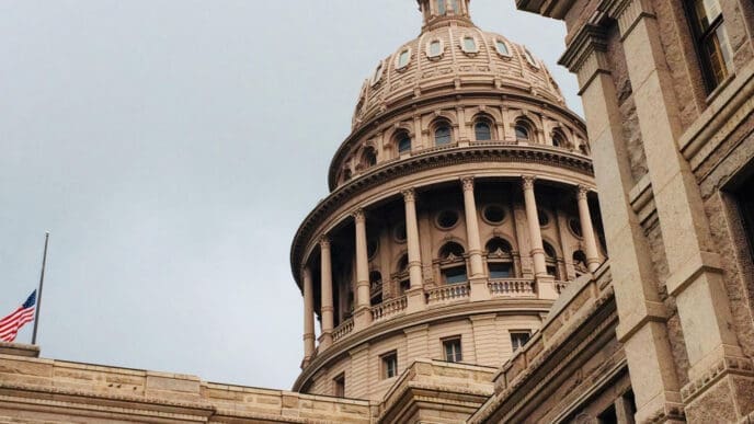 An architectural shot of the Texas State Capitol building, featuring its grand dome and columns, with a US flag at half-staff in the foreground, under an overcast sky.