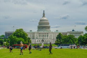 The US Capitol Building with its dome is visible in the background, with a large green lawn in the foreground where people are playing and walking.