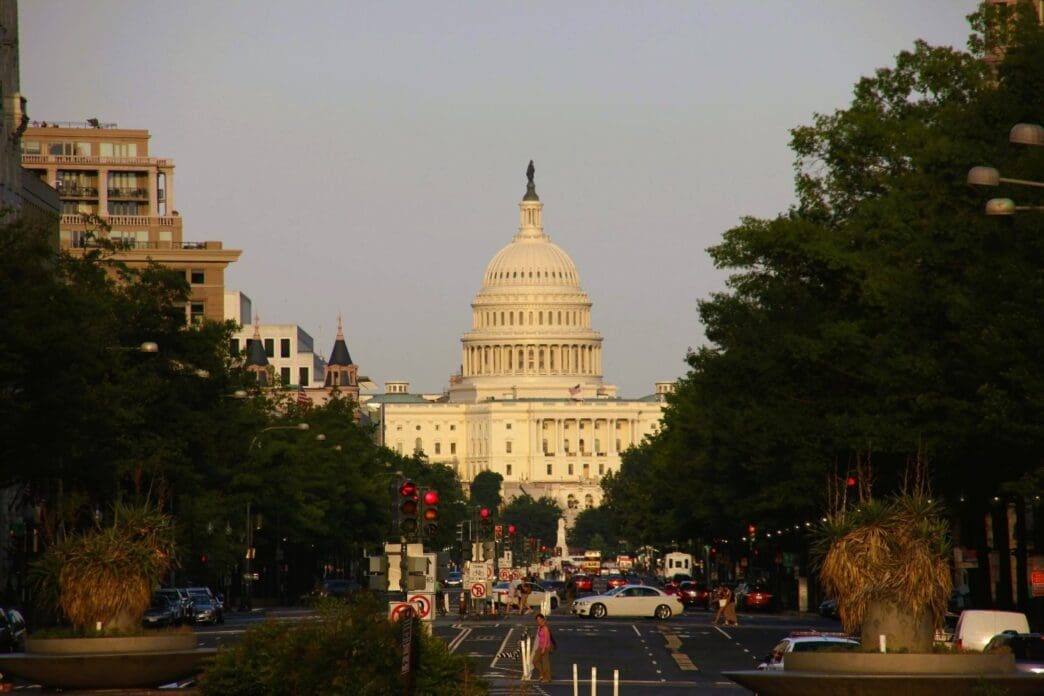 A street view of the US Capitol Building with its white dome, visible at the end of a long, tree-lined avenue with traffic.