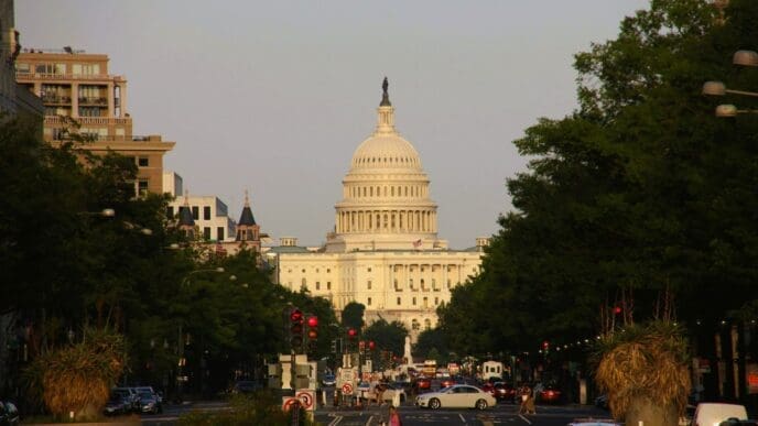 A street view of the US Capitol Building with its white dome, visible at the end of a long, tree-lined avenue with traffic.