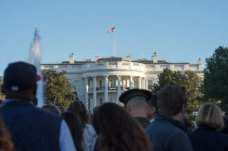 A crowd of people looks toward the back of the White House, with a fountain in the foreground and the American flag flying on the roof.