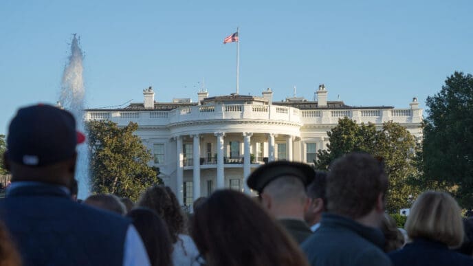 A crowd of people looks toward the back of the White House, with a fountain in the foreground and the American flag flying on the roof.