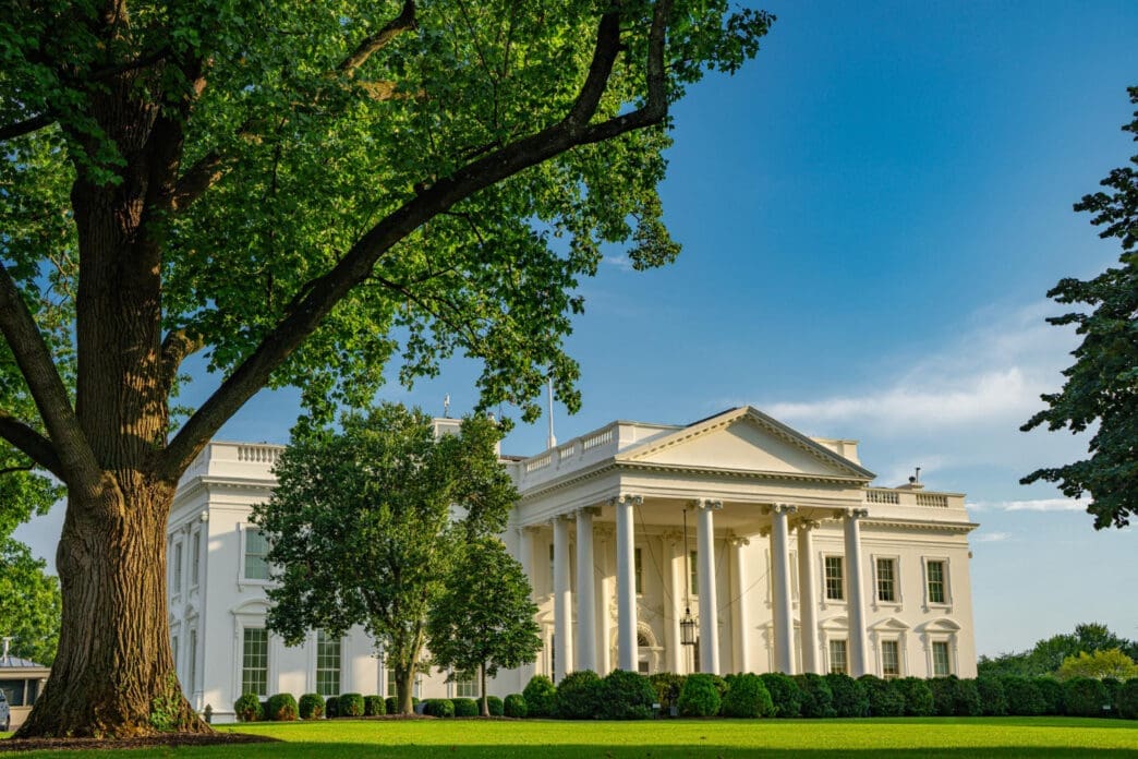 The White House is visible on a sunny day with lush green trees in the foreground.
