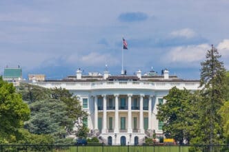 The White House is photographed from the rear, showing its distinctive semicircular facade, columns, and a large American flag flying from the roof.
