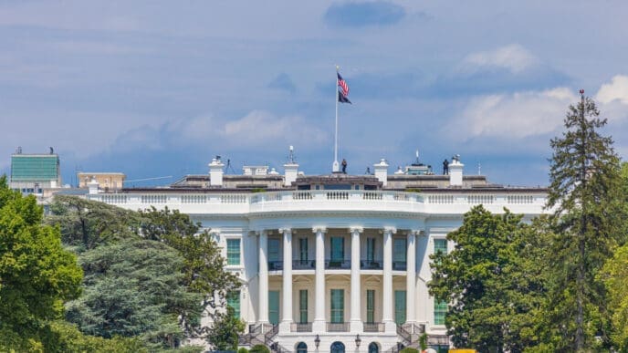 The White House is photographed from the rear, showing its distinctive semicircular facade, columns, and a large American flag flying from the roof.