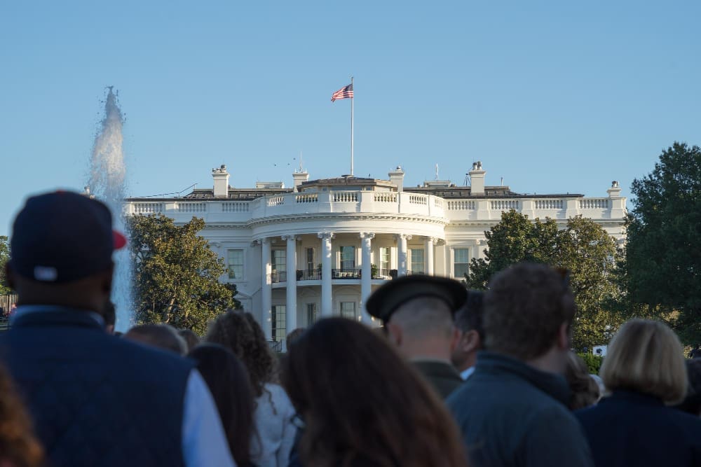 A crowd of people looks toward the back of the White House, with a fountain in the foreground and the American flag flying on the roof.