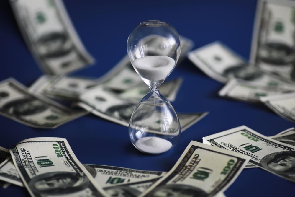 A clear glass hourglass stands on a blue surface, surrounded by scattered one-hundred dollar bills.