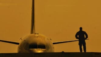 The silhouette of a person stands on a platform in front of the nose and wings of a commercial airplane against a golden sunset sky.