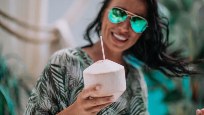 A smiling woman with reflective sunglasses holds a young coconut with a straw in a tropical setting.