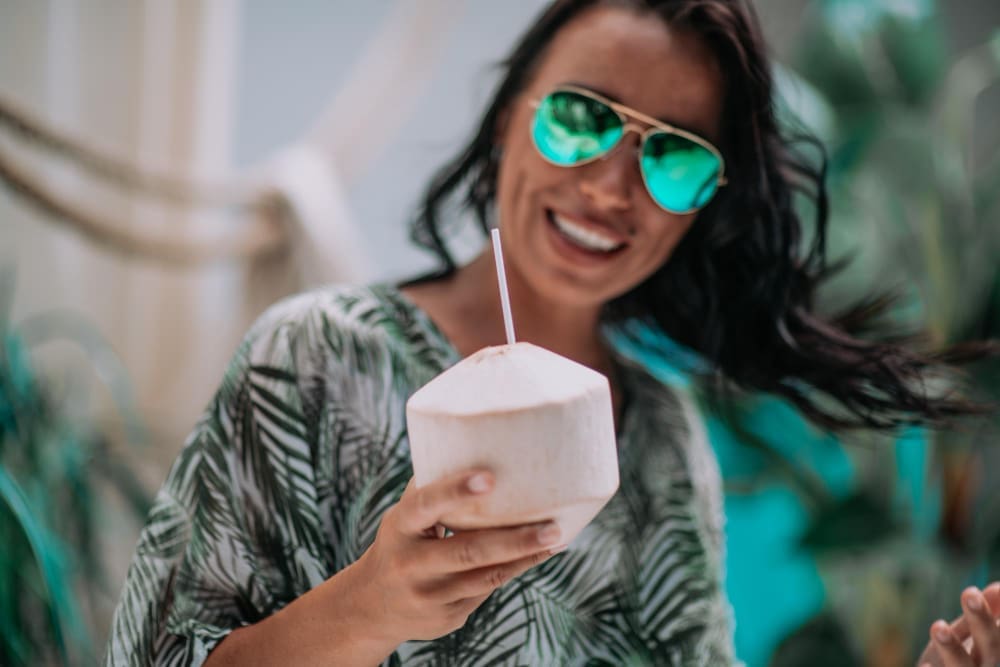 A smiling woman with reflective sunglasses holds a young coconut with a straw in a tropical setting.