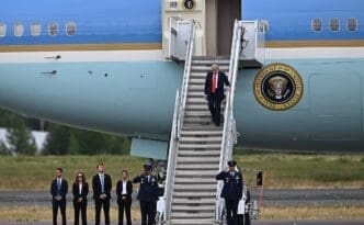 President Donald Trump disembarks from Air Force One, walking down the stairs past a line of officials and military personnel in Alaska.