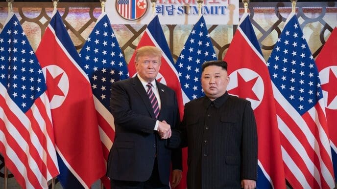 President Donald Trump and North Korean leader Kim Jong Un are standing and shaking hands in front of a row of American and North Korean flags at a summit.